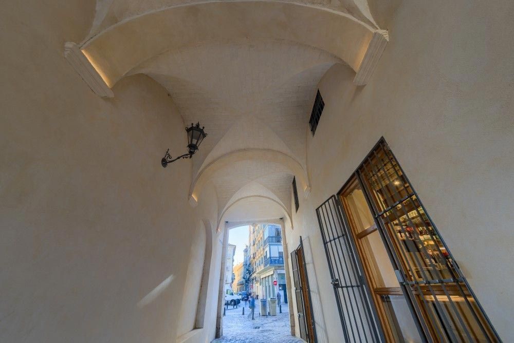 Un estrecho pasadizo de piedra con arcos desemboca en una calle adoquinada iluminada por el sol, con edificios azules al fondo.