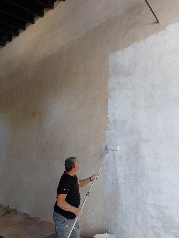 Persona pintando una pared grande con un rodillo en un palo largo en un espacio interior.