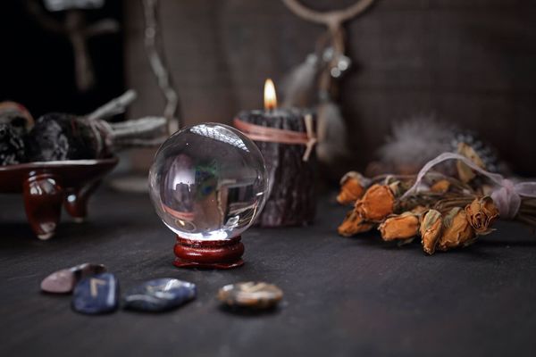 Bola de cristal sobre un soporte de madera, vela encendida y runas sobre una superficie oscura, lo que sugiere adivinación.