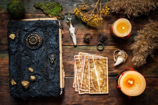 Escena de brujería: un libro de hechizos, cartas del tarot, velas, anillos, cristales y hierbas secas sobre madera.