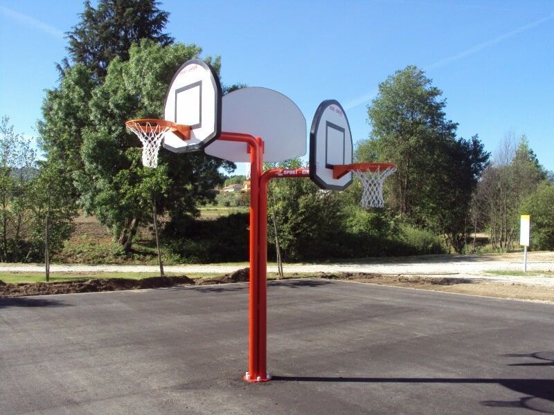 Uma estrutura de basquete com postes vermelhos, duas tabelas e aros posicionados em direções opostas em uma quadra ar livre