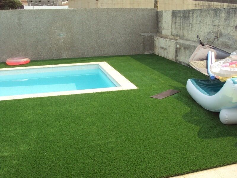 Uma pequena piscina retangular rodeada por relva artificial verde, com boias e uma espreguiçadeira por perto.