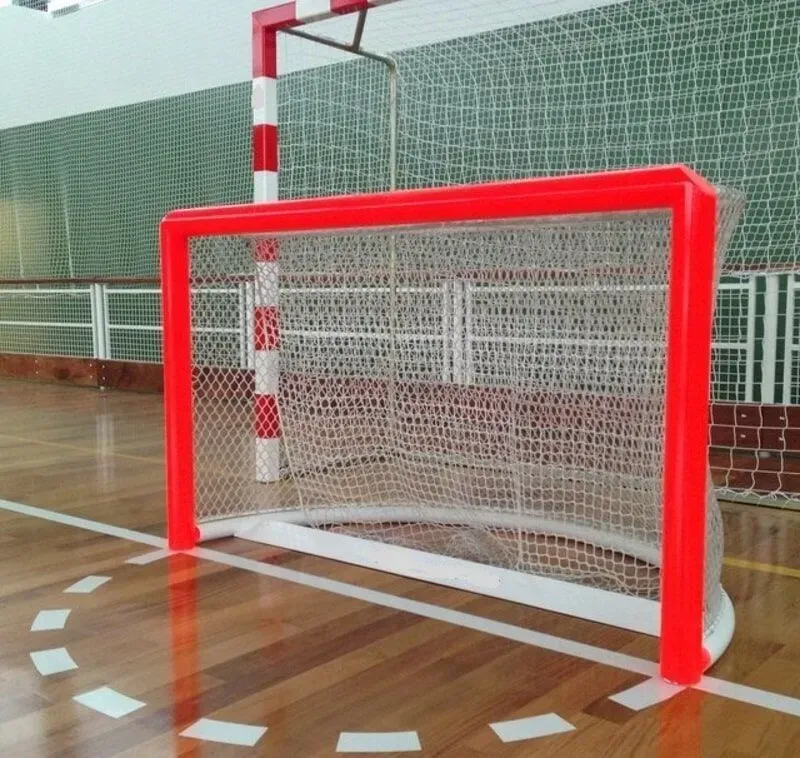 Uma baliza de handebol indoor vermelha e branca com rede está localizada em uma quadra de madeira com marcações brancas.