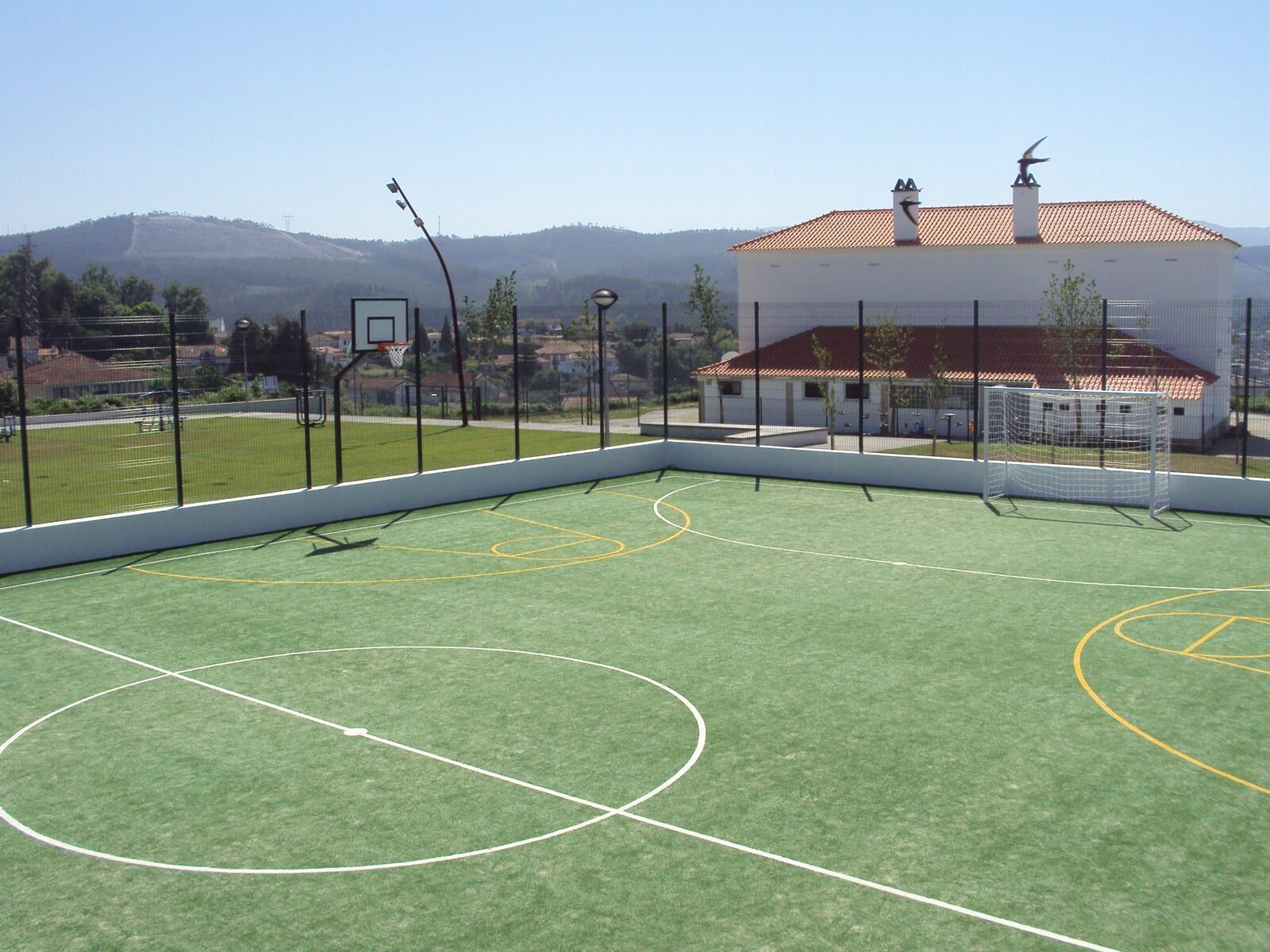 Uma quadra esportiva ao ar livre com grama verde, linhas de demarcação brancas, cestas de basquete e um prédio