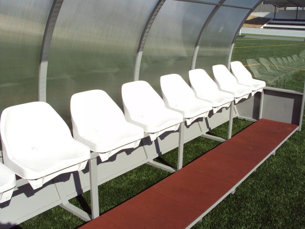 Uma fileira de assentos brancos de estádio sob uma cobertura curva transparente em um campo esportivo.