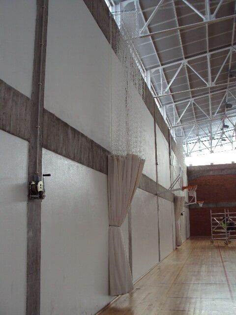 Interior de ginásio, vigas horizontais escuras e uma cortina parcialmente fechada que vai do chão ao teto.