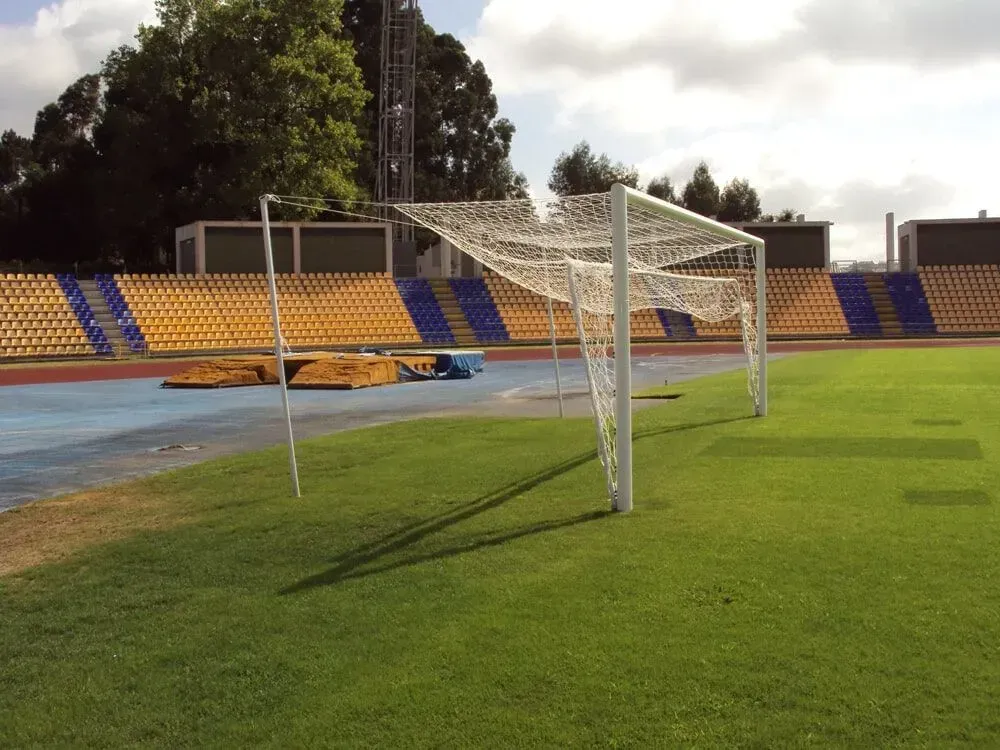 Um gol de futebol com redes brancas está de pé em um campo gramado em um estádio vazio com arquibancadas amarelas e azuis.