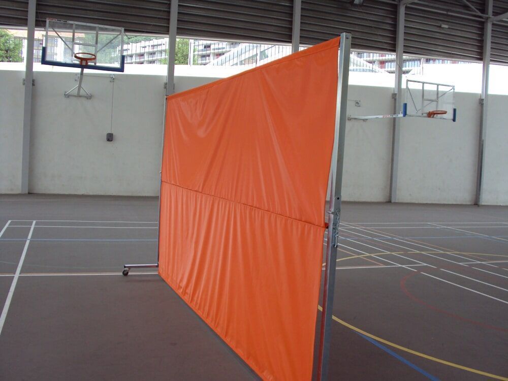 Uma divisória portátil laranja está posicionada em uma quadra de basquete coberta, em frente a uma cesta.