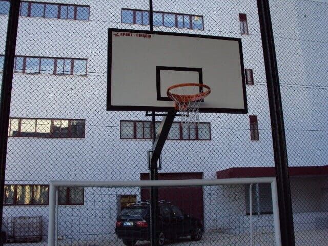 Uma cesta de basquete montada em um poste com uma tabela, vista através de uma cerca de tela metálica com um prédio ao fundo.