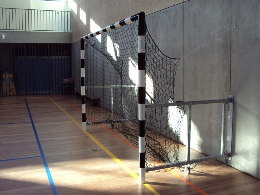 Uma baliza de andebol com postes listrados em preto e branco, fixada numa parede de concreto em um ginásio coberto.