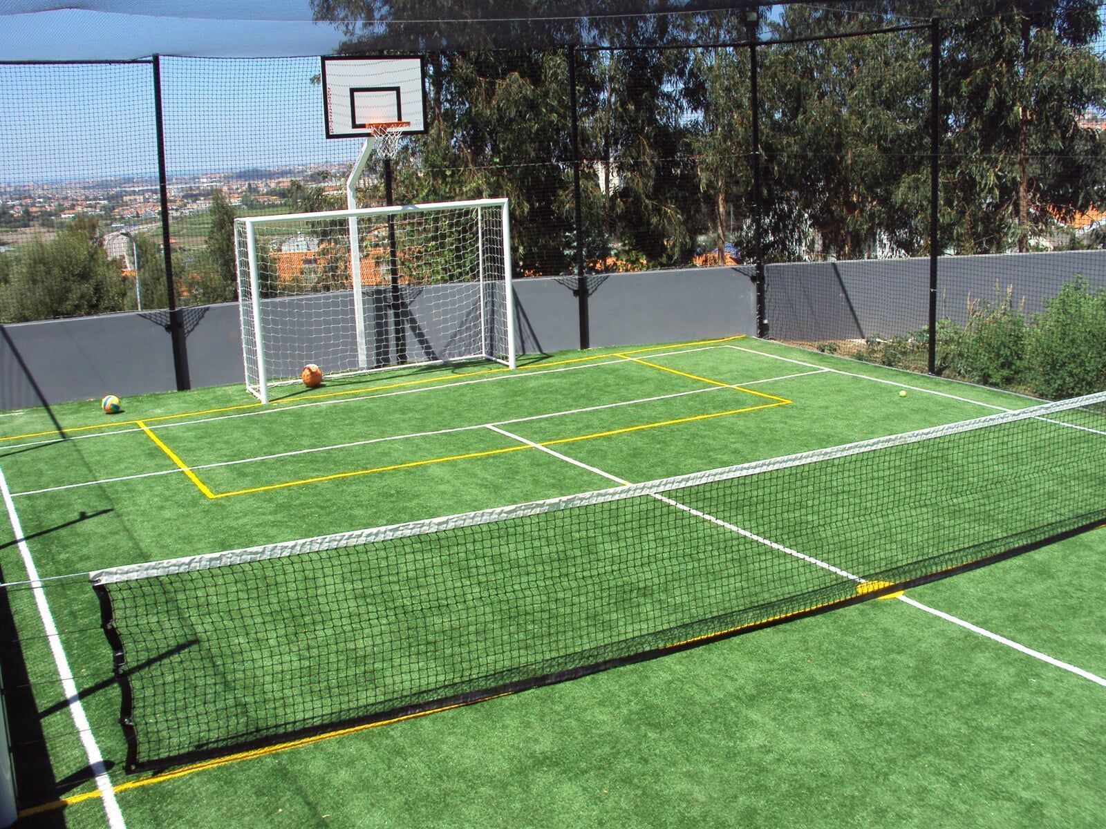 Uma quadra poliesportiva de grama sintética com rede de tênis, gol de futebol e cesta de basquete, cercada por uma rede alta.