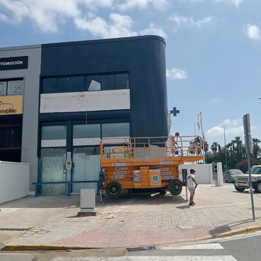 Una plataforma elevadora de tijera naranja y un trabajador frente a un edificio oscuro en construcción, en una esquina soleada