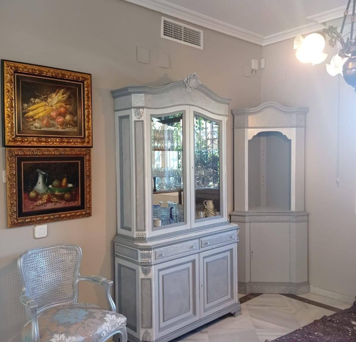 Elegante rincón de salón con vitrina blanca, cuadros enmarcados, silla plateada y suelo de mármol.