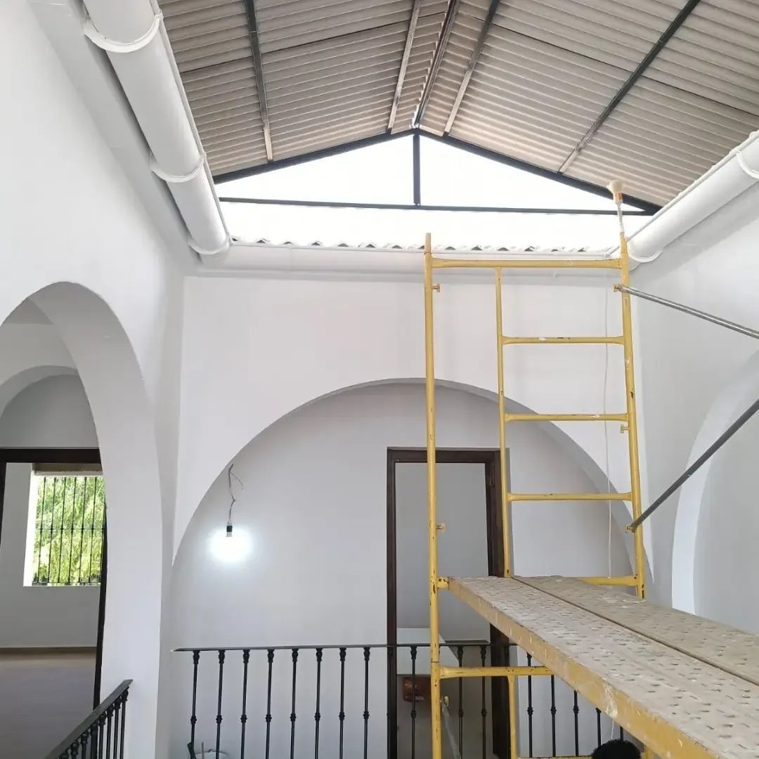 Interior con paredes blancas arqueadas, techo a la vista y un andamio amarillo sobre una pasarela de entreplanta.
