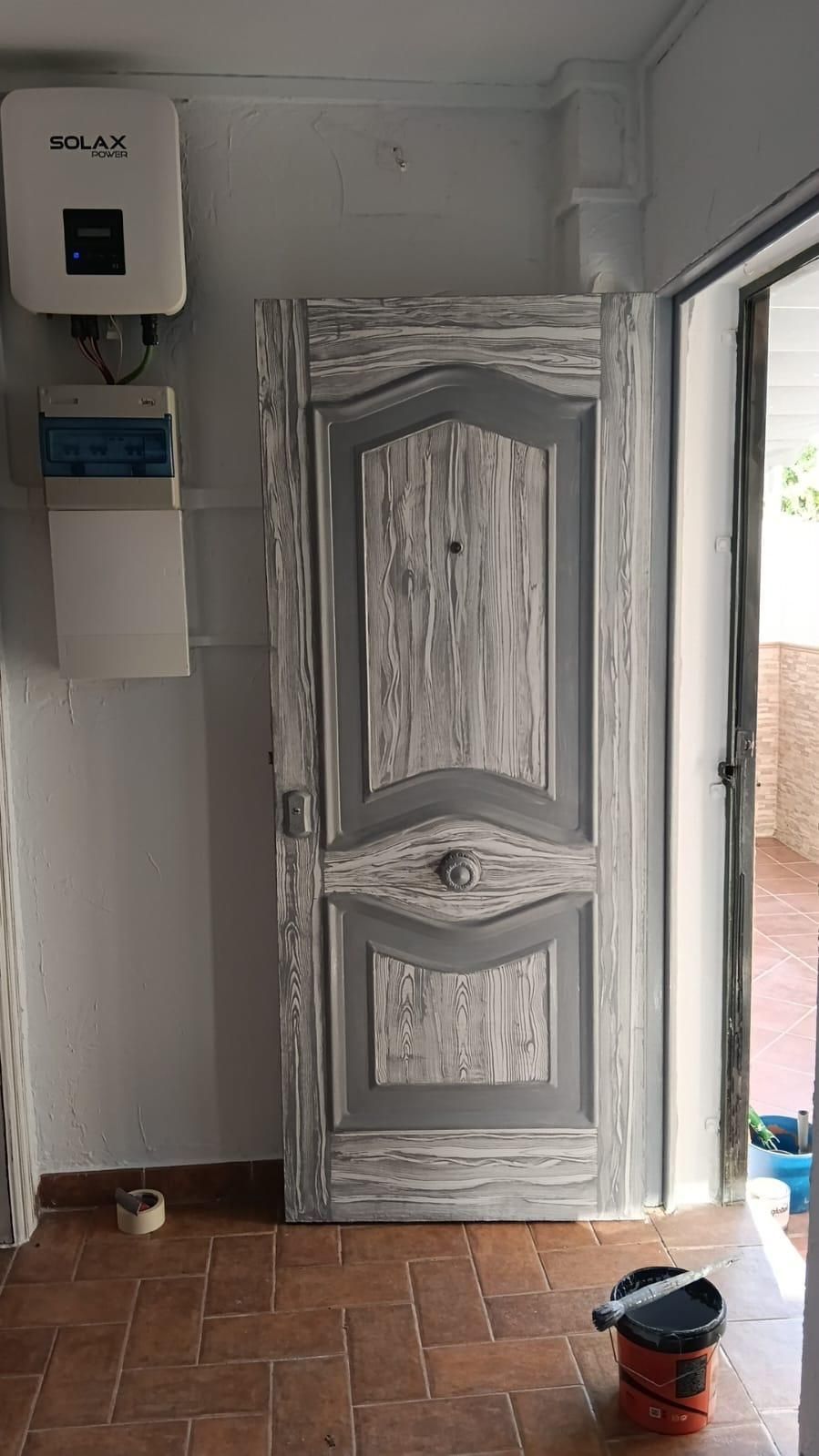 Cocina pequeña con una puerta de madera tallada de color gris, armarios de pared blancos y suelo de baldosas; escoba y cubo cerca de la entrada