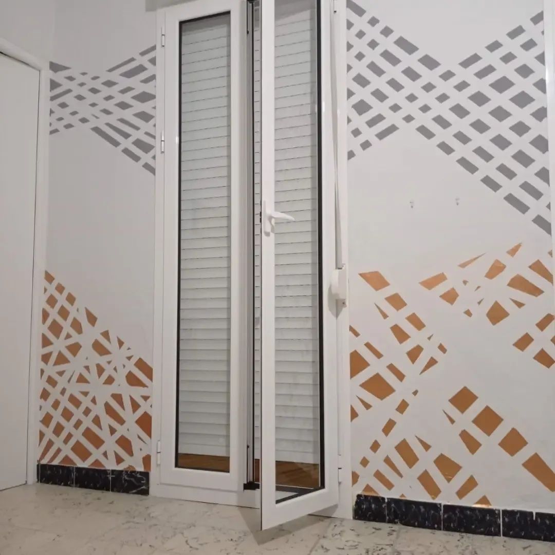 Puerta exterior blanca con paneles de vidrio y adhesivos geométricos grises y naranjas en la pared.