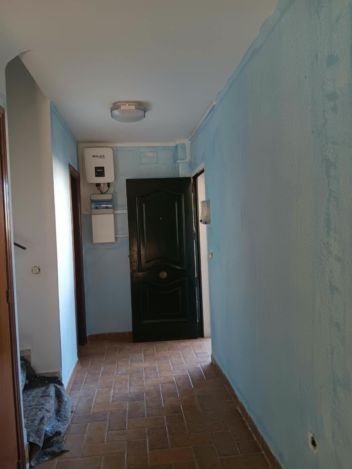 Pasillo estrecho azul con una puerta verde oscuro, suelo de baldosas de terracota y una luz en el techo.