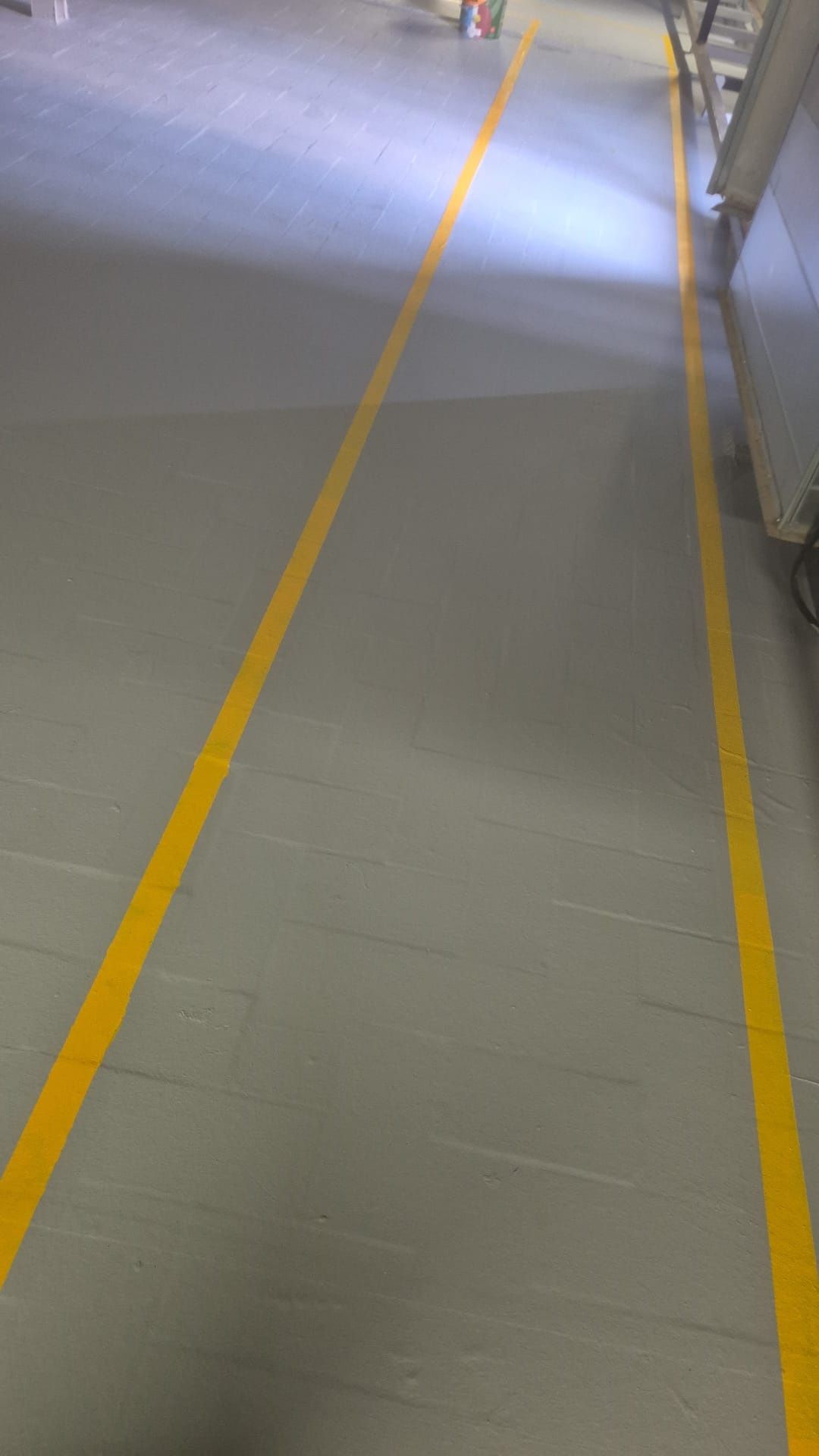 Pasillo interior con suelo gris y dos líneas guía amarillas que conducen a una puerta iluminada.