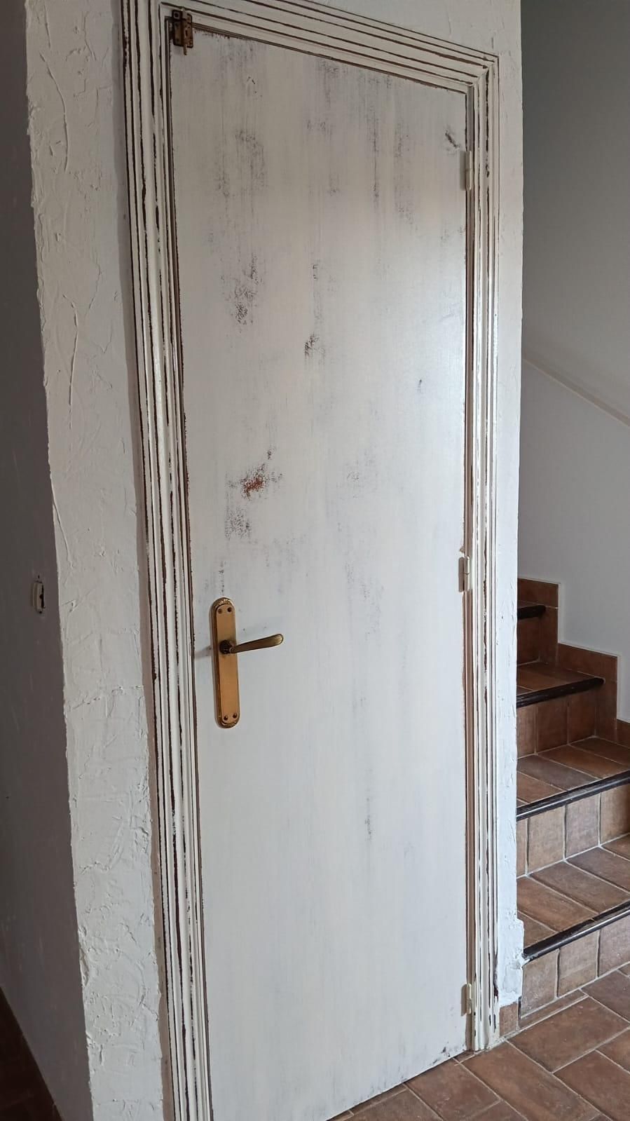 Puerta blanca desgastada con tirador de latón junto a una escalera en el interior de una vivienda