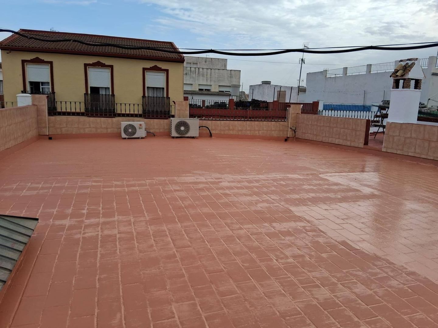 Terraza en la azotea con baldosas de terracota, muros bajos de ladrillo y edificios vecinos bajo un cielo nublado