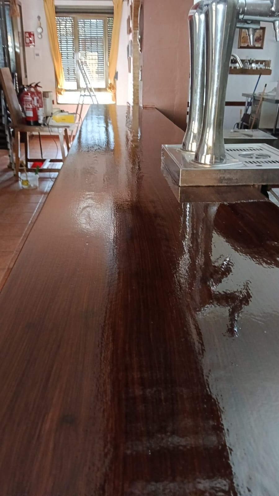 Una larga mesa de madera en un comedor luminoso, con un tubo metálico de estufa y sillas junto a una ventana.
