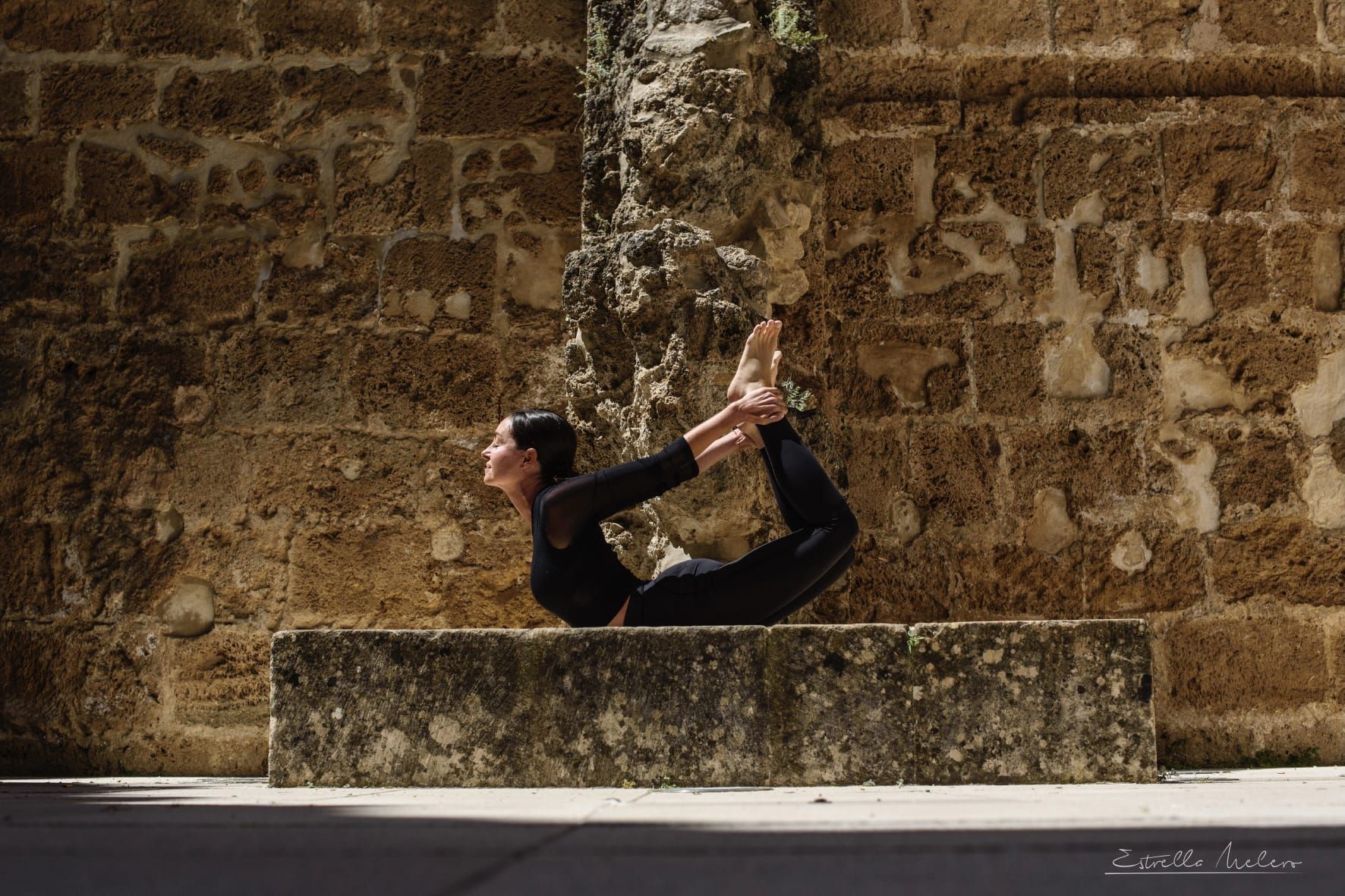 Una persona realiza una postura de yoga sobre una repisa de piedra, apoyada contra una pared de ladrillo texturizado.
