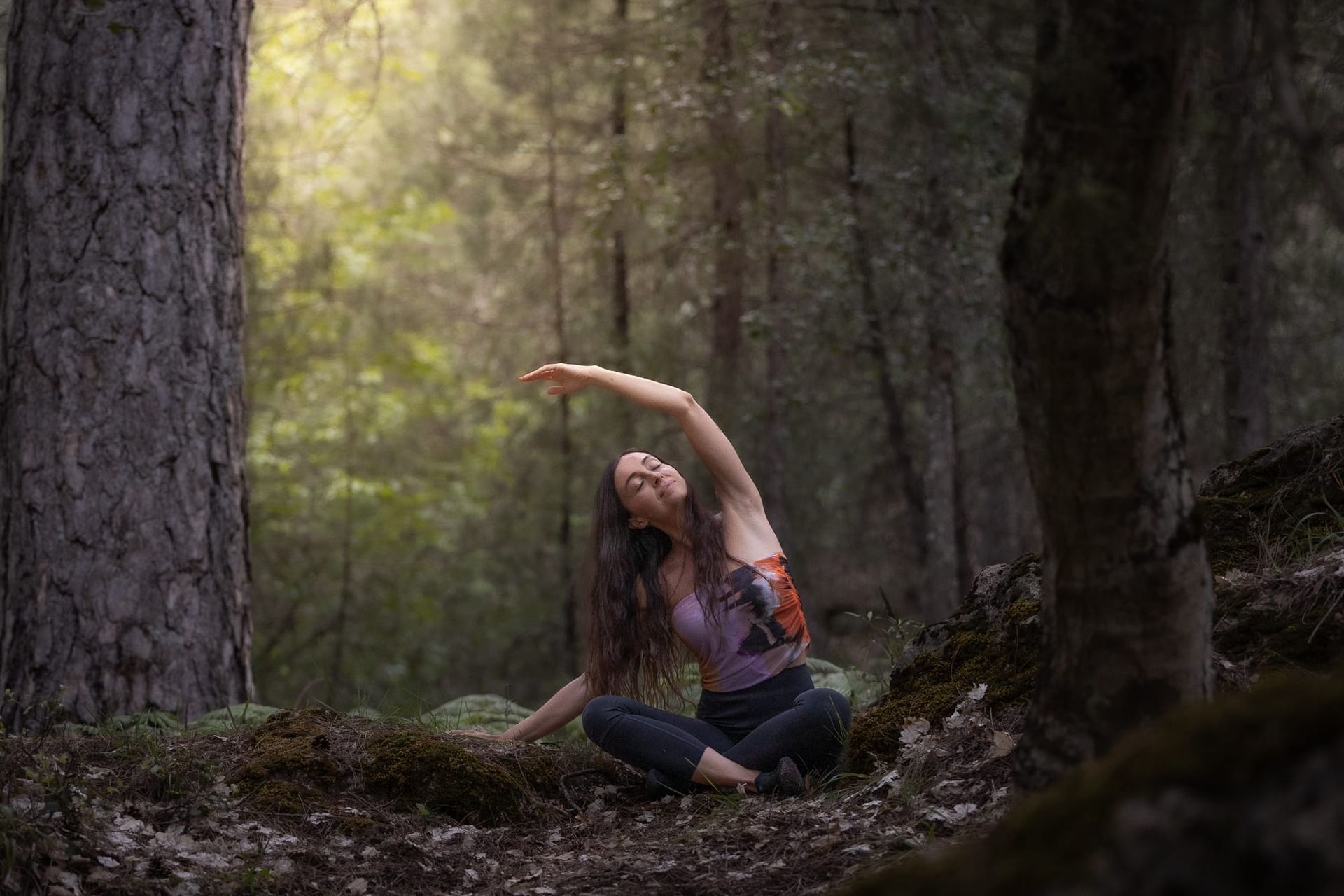 Mujer estirándose en un bosque, sentada en un sendero cubierto de musgo con los brazos en alto bajo la luz del sol moteada.