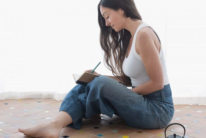 Woman sitting on the floor, writing in a notebook beside a window.