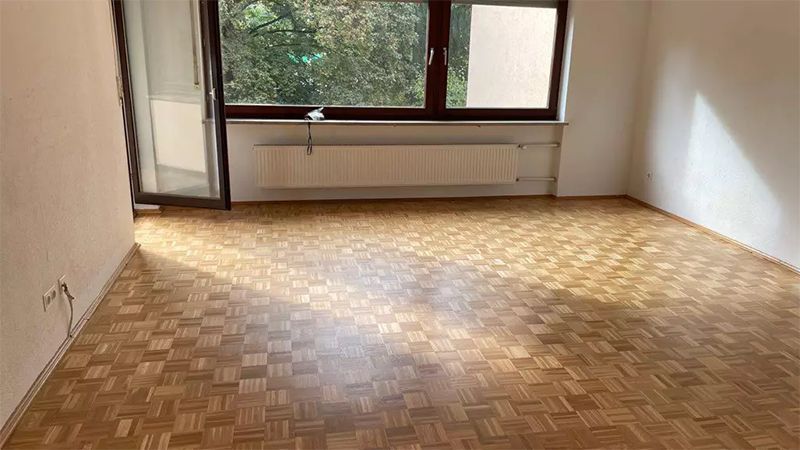Leeres Zimmer mit Parkettboden, einem Fenster und einer Tür zu einem Balkon. Sonnenlicht durchflutet den Boden.