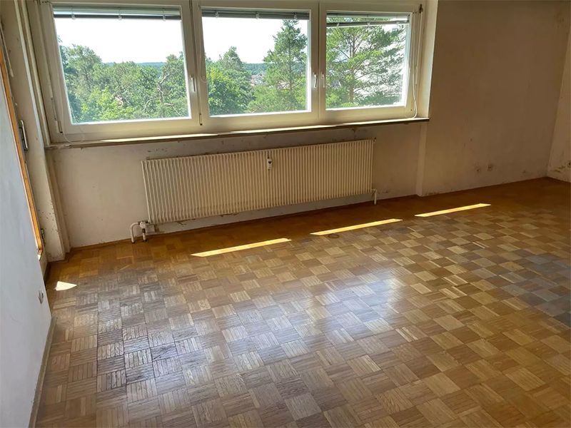 Leeres Zimmer mit kariertem Holzboden, Heizkörper und einem dreiteiligen Fenster mit Blick auf Bäume.