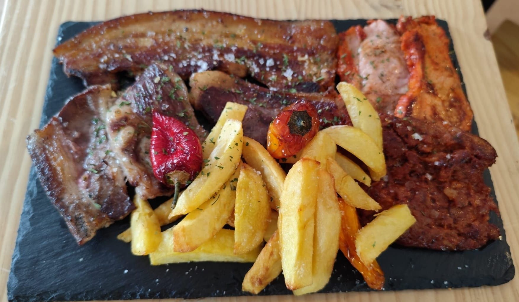 Surtido de carnes a la parrilla y patatas fritas sobre pizarra negra, con un pequeño pimiento.