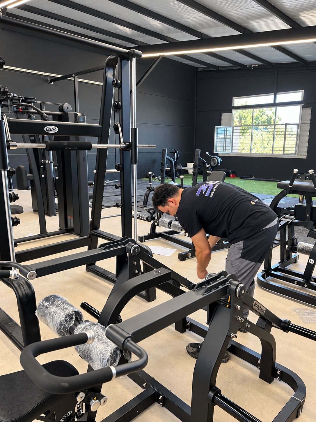 Persona inclinada sobre una máquina de gimnasio en una sala de musculación moderna, ajustando el equipo