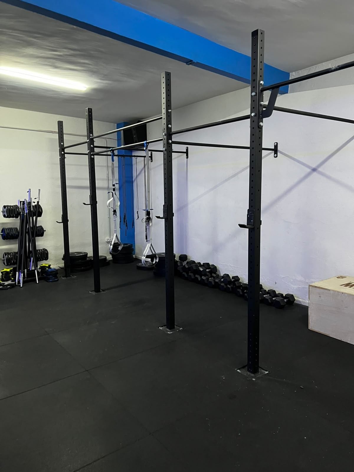 Zona de gimnasio vacía con una jaula de sentadillas, mancuernas y suelo negro bajo luces cenitales