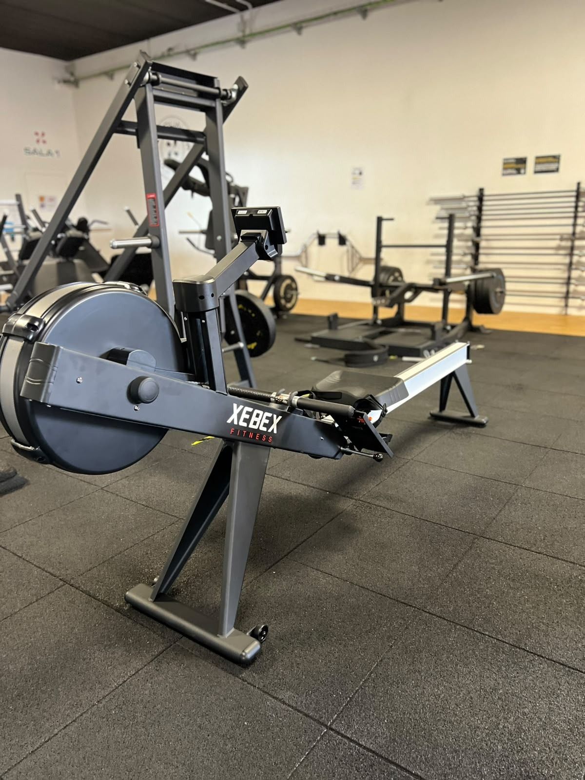 Máquina de remo en un gimnasio, con estructura negra y un gran volante de inercia sobre un suelo de goma.