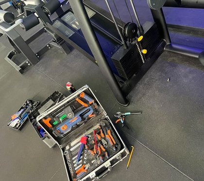 Caja de herramientas abierta con herramientas manuales en el suelo de un gimnasio, junto a equipos de ejercicio

