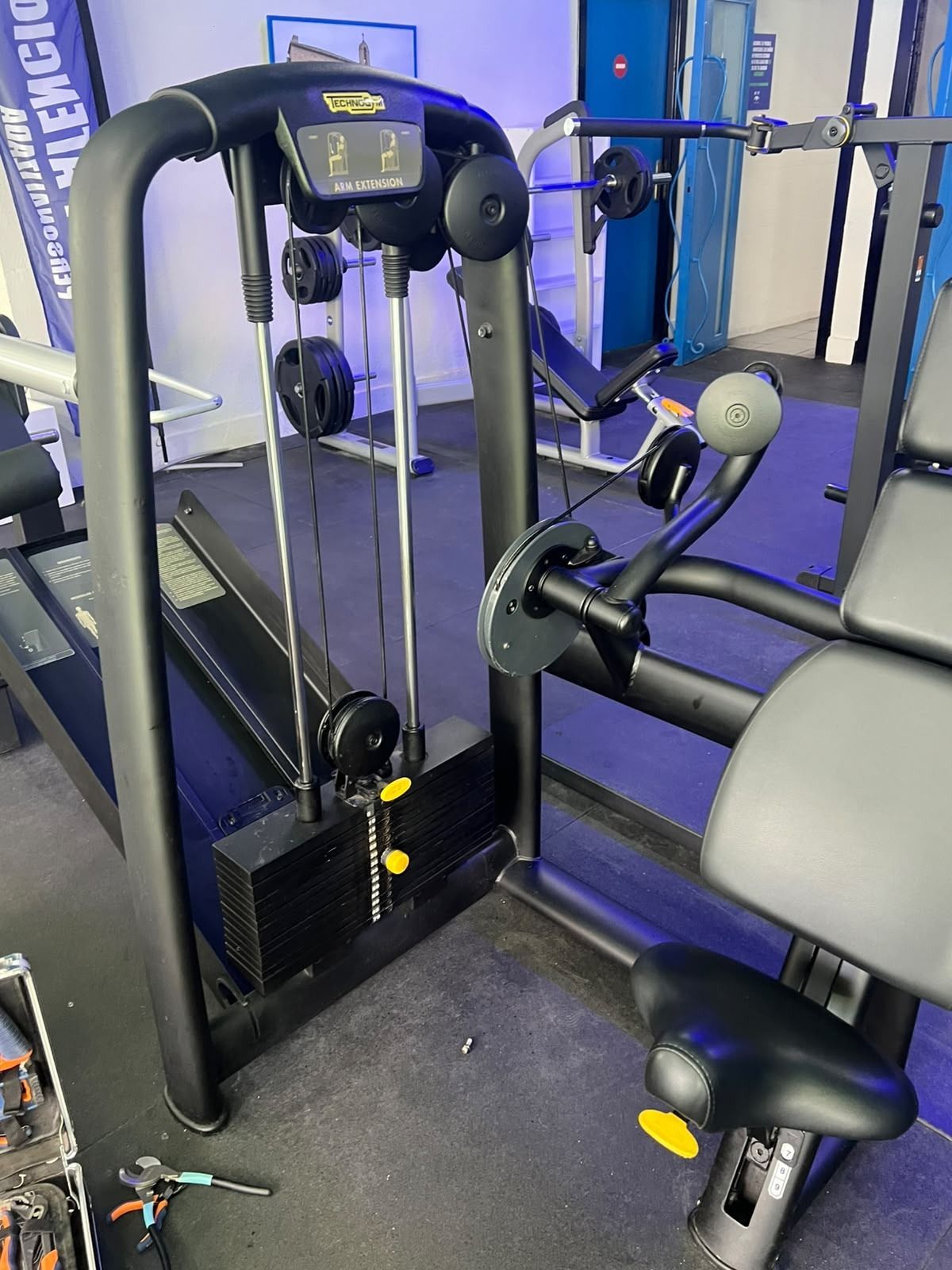 Máquina de gimnasio con asientos y empuñaduras acolchados en negro y gris, en una sala de fitness con paredes azules