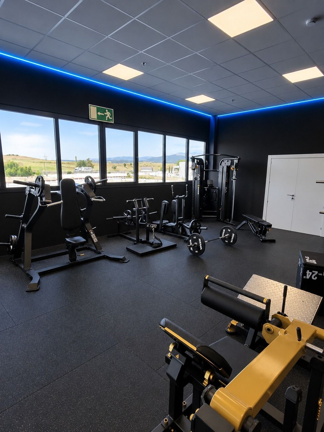 Interior de un gimnasio vacío, con máquinas de ejercicio, suelo negro, grandes ventanales y luces LED azules en el techo