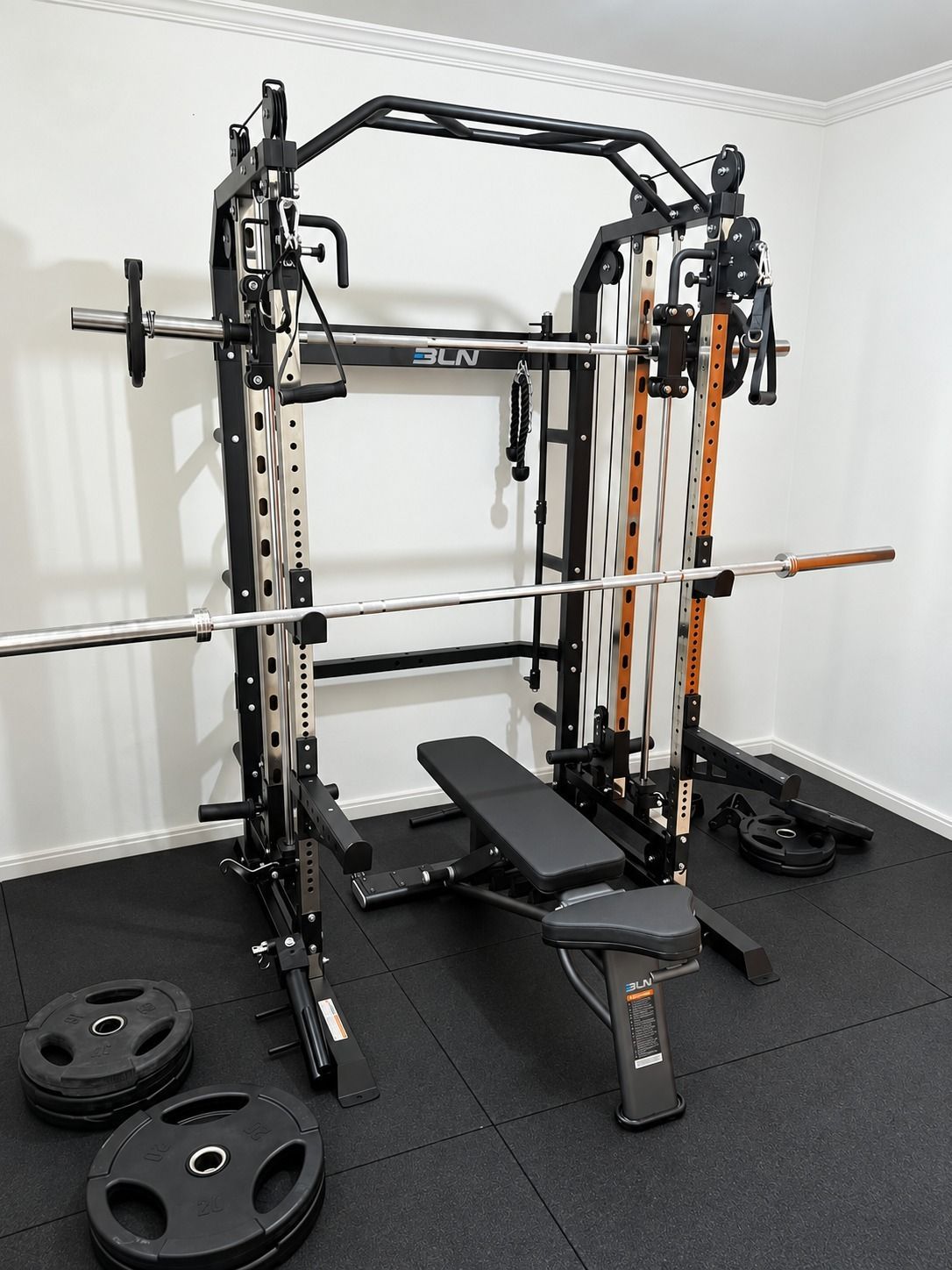 Gimnasio en casa con una jaula de potencia, un banco, una barra y discos de pesas en una habitación pequeña.