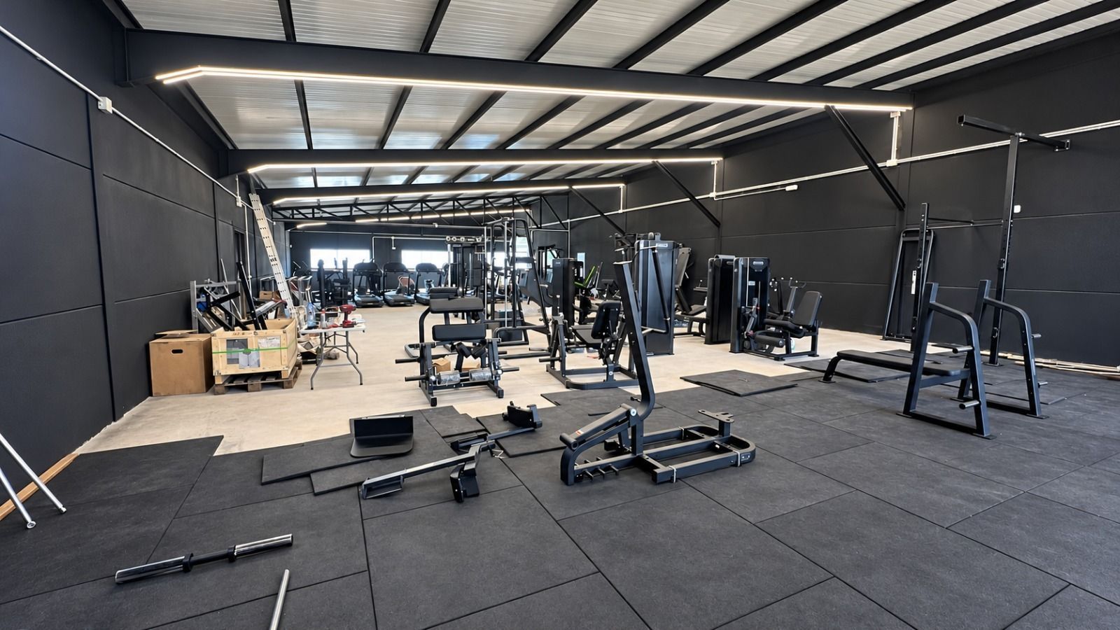 Gimnasio vacío con suelo negro, máquinas de pesas, bancos y grandes lámparas industriales en el techo.