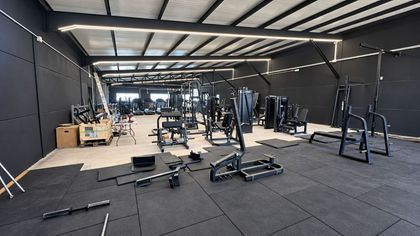 Gimnasio vacío con suelo negro, máquinas de pesas, bancos y grandes lámparas industriales en el techo.