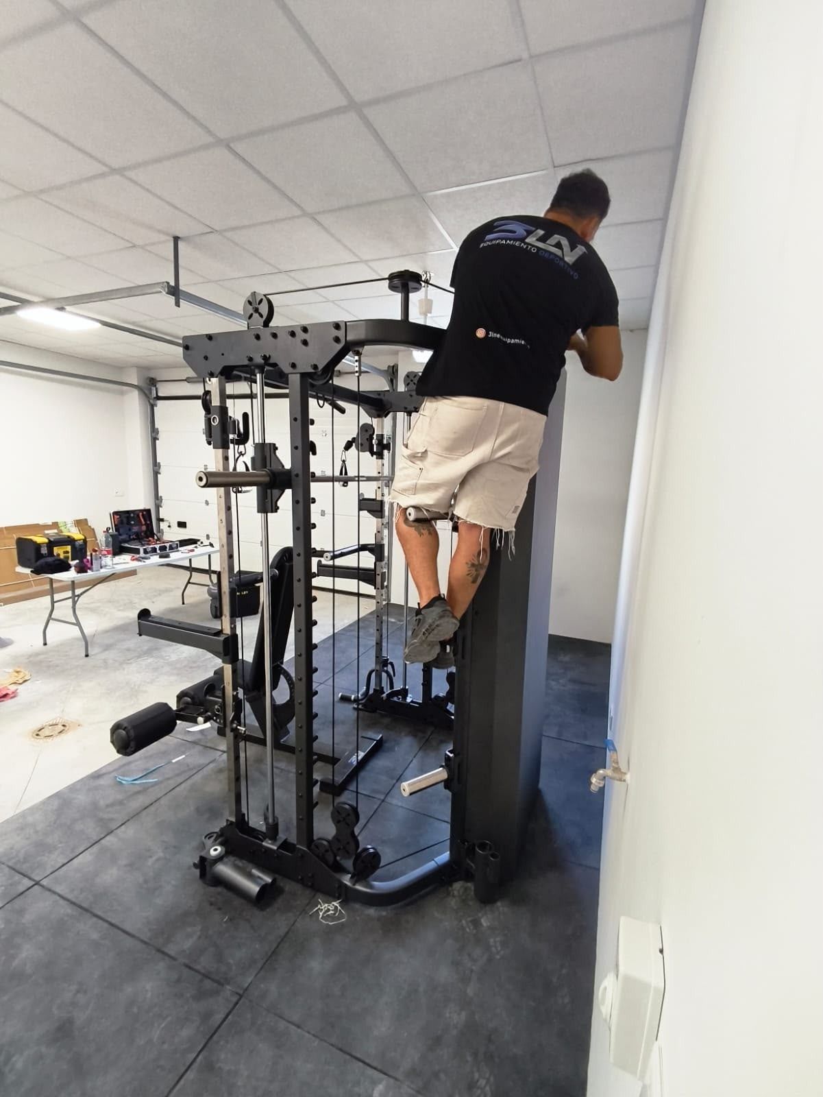 Persona ajustando una máquina de poleas en una sala de gimnasio con suelo de goma negro y paredes blancas