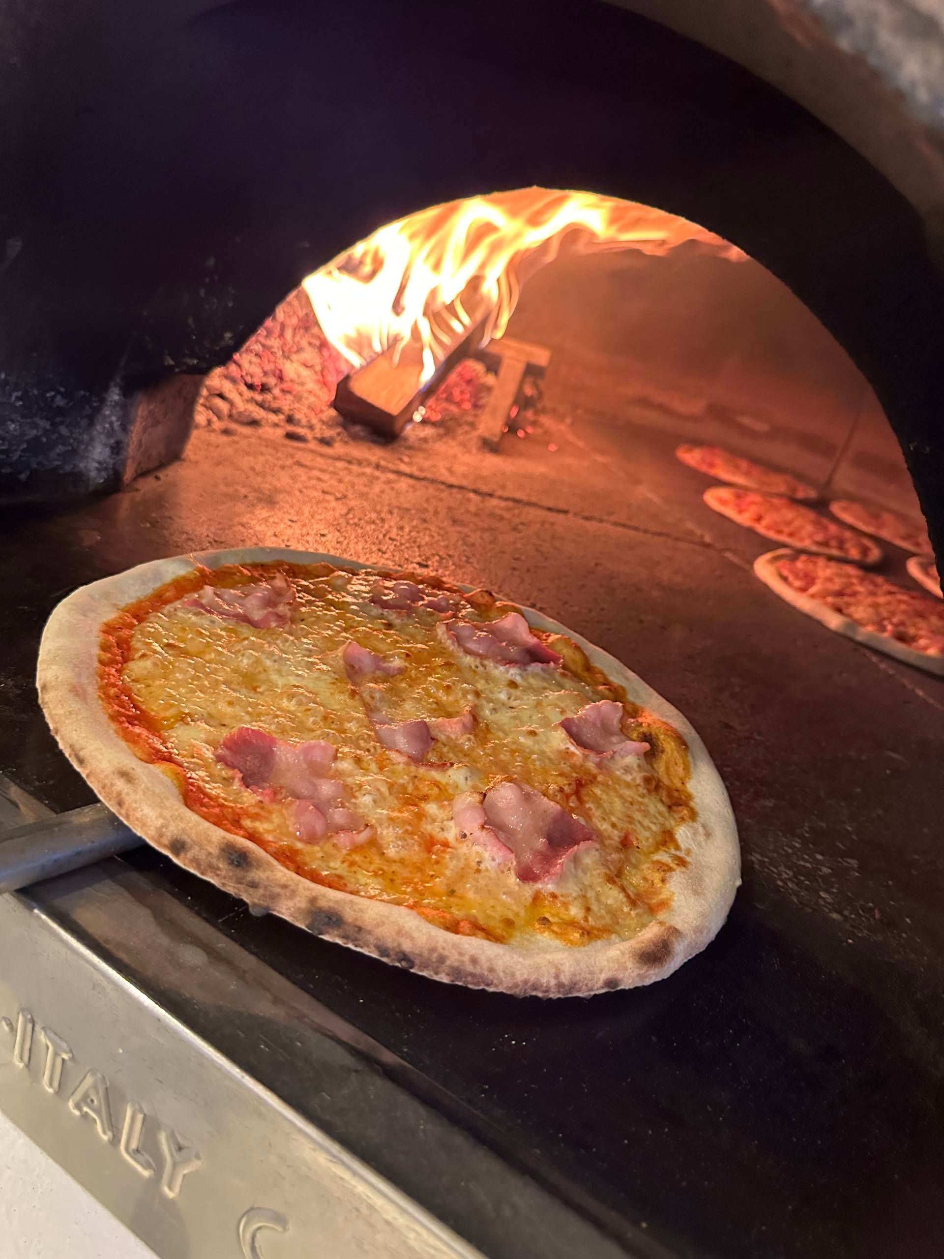 Pizza, die in einem Holzofen mit sichtbaren Flammen gebacken wird; Nahaufnahme einer Pizza auf einem Pizzaschieber.
