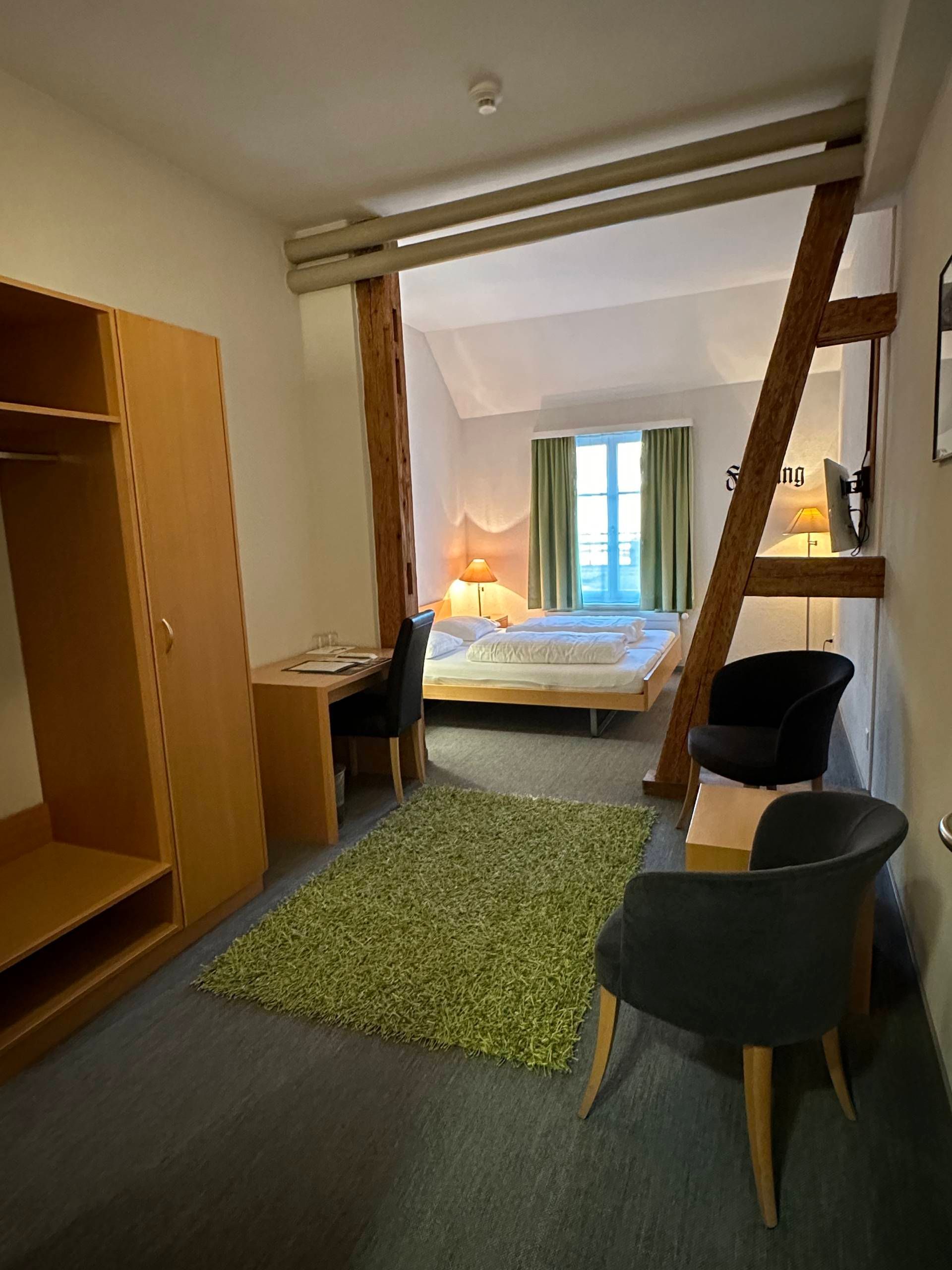 Hotelzimmer mit Bett, Schreibtisch, Kleiderschrank, Stühlen und grünem Teppich.