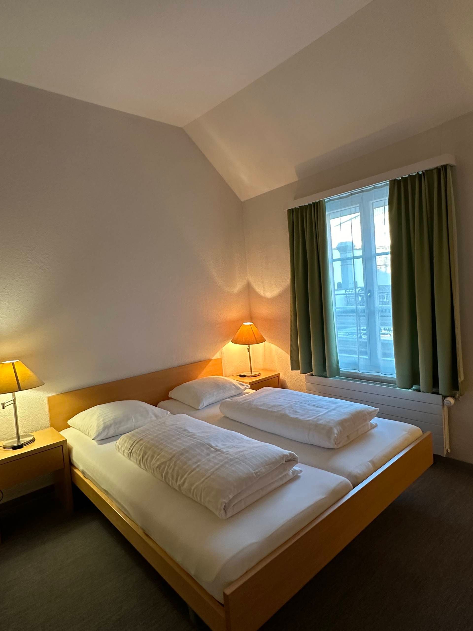 Schlafzimmer mit zwei Einzelbetten, kleinen Nachttischen und Fenster mit grünen Vorhängen.