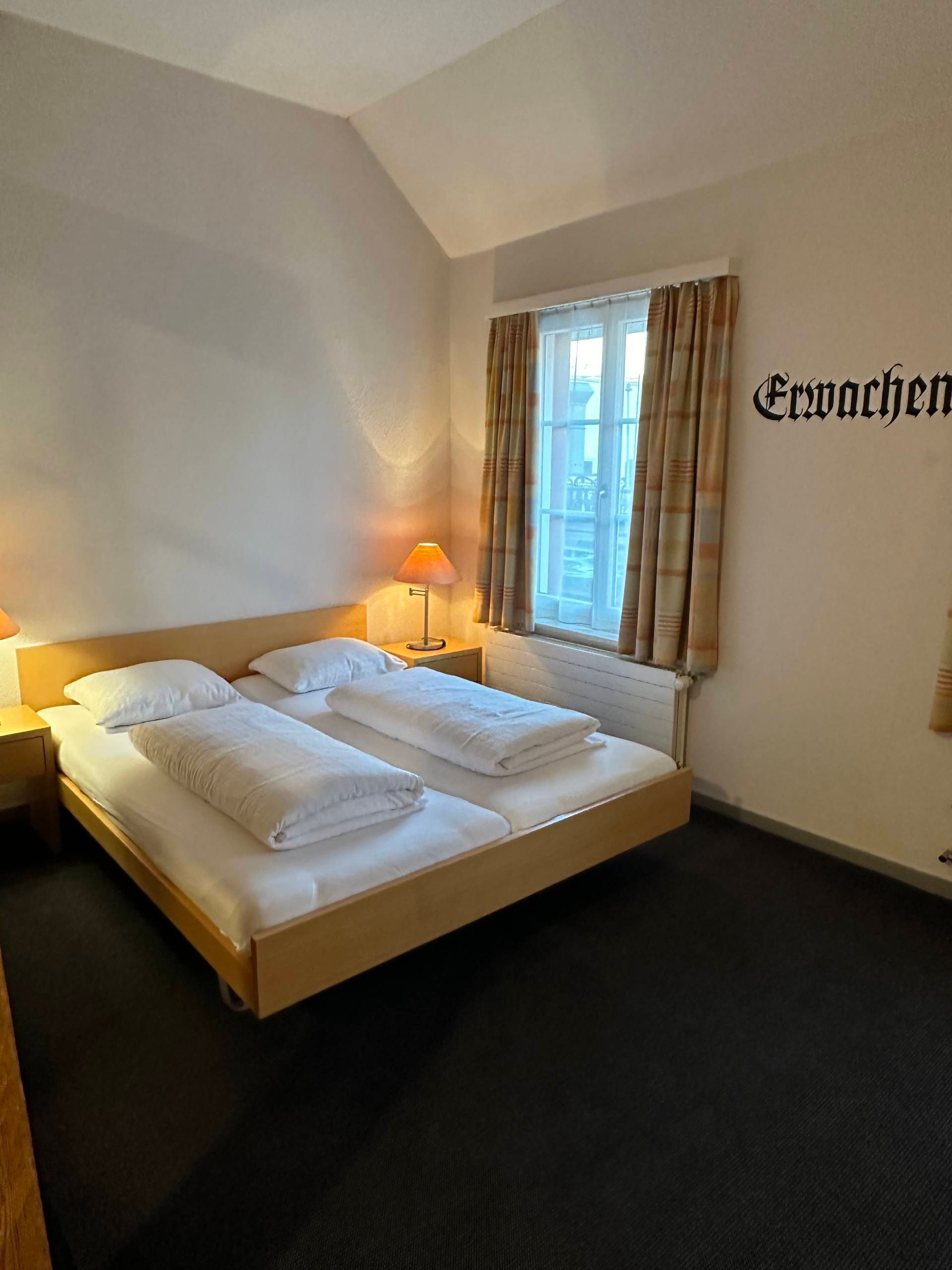 Schlafzimmer mit Doppelbett, Nachttischen, einem Fenster und dem Wort „Erwachen“ an der Wand.