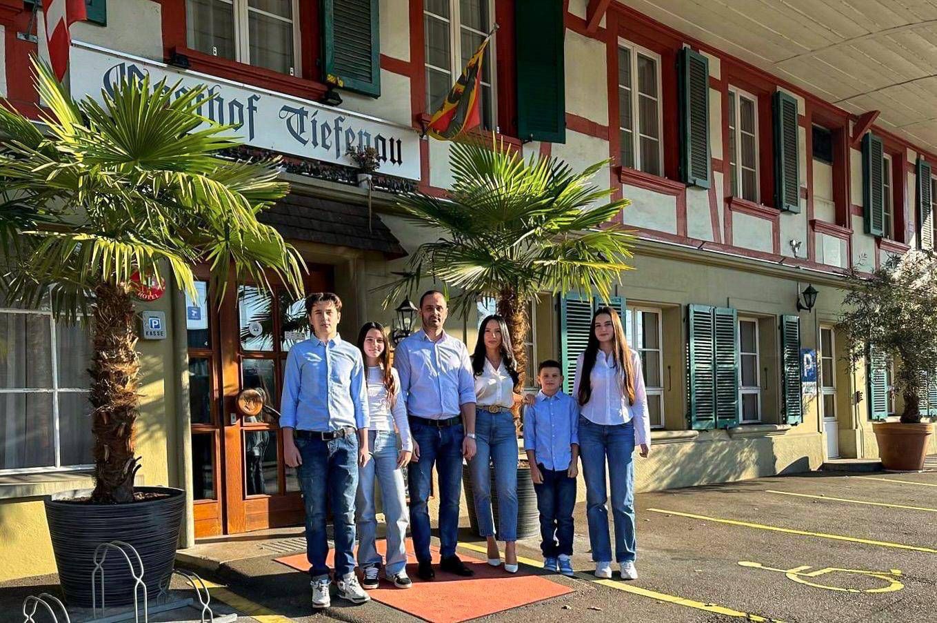 Eine Familie posiert vor einem Restaurant. Das Gebäude hat eine rot-weisse Fassade. Palmen säumen den Eingang.