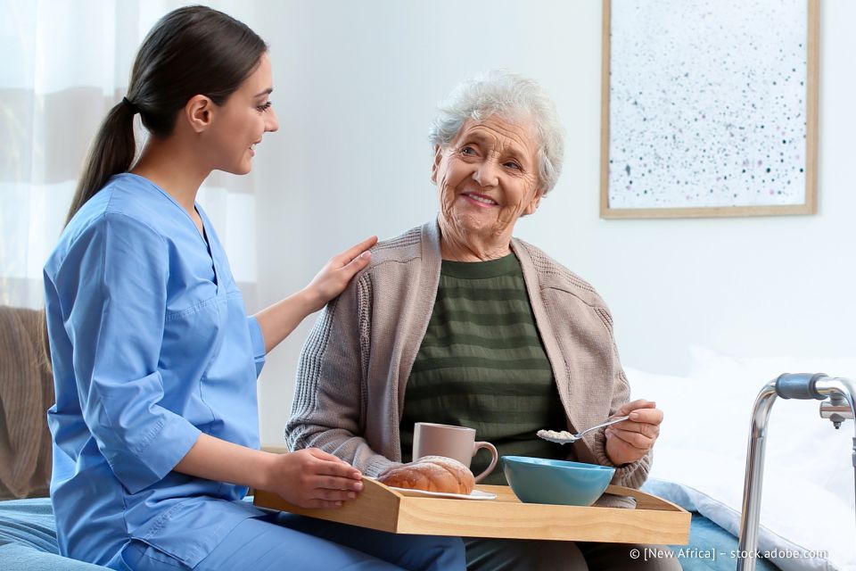 Pflegekraft in blauer OP-Kleidung mit der Hand auf der Schulter einer älteren Frau, die lächelt und in einem Schlafzimmer von einem Tablett isst.