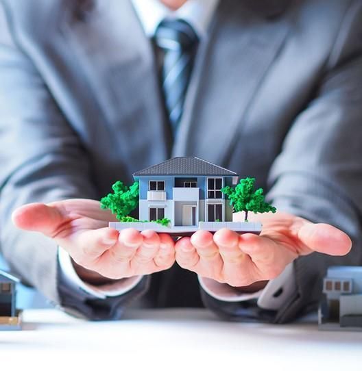 Une personne en costume tient une maison miniature, peut-être une maquette ou une représentation architecturale.