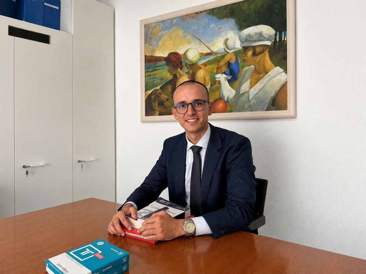 Un hombre con traje azul marino sonríe junto a una mesa de conferencias con folletos, en una oficina con un gran cuadro detrás de él.