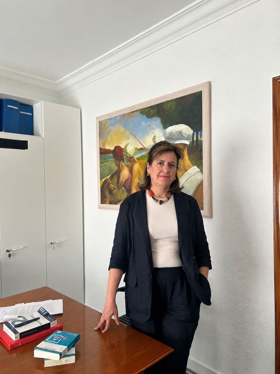 Persona de pie junto a un escritorio en una oficina luminosa, con un cuadro en la pared y libros sobre la mesa.